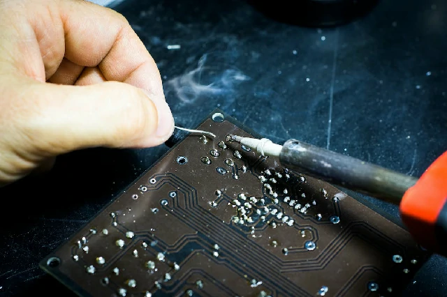 Un técnico soldando cuidadosamente componentes en una placa de circuito impreso. El humo se eleva desde el punto de contacto donde el soldador aplica el estaño, indicando la precisión del trabajo manual en la fabricación de hardware electrónico.