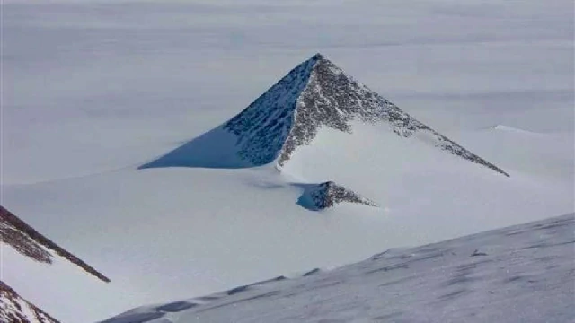 Imagen de una montaña con forma piramidal cubierta parcialmente de nieve, emergiendo del paisaje helado de la Antártida. La simetría y la pendiente pronunciada de sus lados contrastan con el vasto y plano entorno nevado.