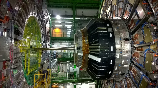 La imagen muestra el interior de una instalación del CERN, con un gran detector de partículas en el centro, rodeado de equipos y estructuras metálicas complejas. La maquinaria, con paneles y componentes de diferentes colores, está dispuesta en un entorno industrial iluminado. Esta configuración es típica de los experimentos de física de partículas, donde se realizan investigaciones avanzadas sobre los fundamentos del universo.
