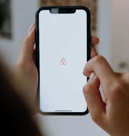 Una persona sostiene un teléfono inteligente con la pantalla mostrando el logotipo de Airbnb sobre un fondo blanco.